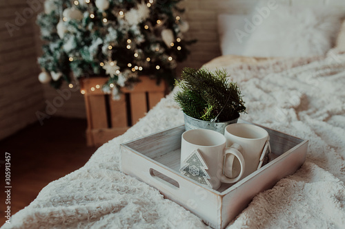 two white mugs on a wooden tray stand on the bed with a fluffy blanket against the background of the tree, a room in a Christmas decor in bright colors and garlands photo