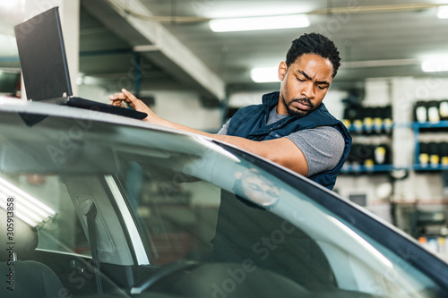 Africa American auto mechanic using laptop while examining car in repair shop.