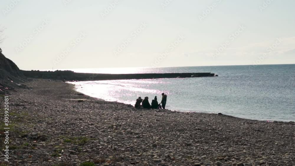 Close-up silhouettes of a family with a dog resting on the beach at sunset in the cold season. Close-up silhouettes of a family with a dog resting on the beach at sunset in the cold season.