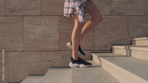 The feet of a young woman in black sneakers, shorts and a plaid shirt climb the stairs, a stone wall in the background. Slow motion. Tracking camera on the object.