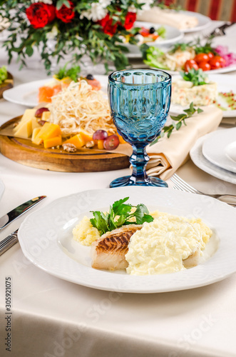 Fried cod white fish with mashed potatoes on a white plate with hot snack, alads, cutlery, wine and water glasses on banquet restaurant table background. European food in a restaurant.