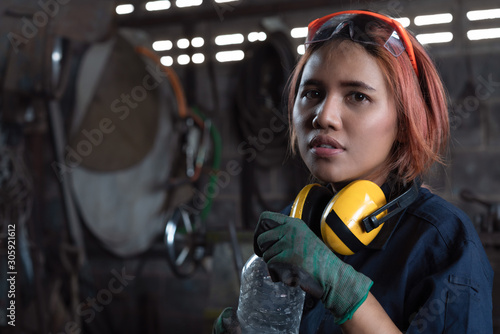 Tired Asian female workshop engineer holding water bottle wearing industrial safety equipment in factory - Young engineering apprentice woman taking break with copy space -Industry and trainee concept