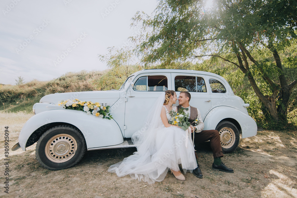 Beautiful newlyweds are sitting and hugging near an old retro car and summer nature. Wedding portrait of a stylish, smiling groom and lovely bride with curly hair. Photography and concept. Beautiful newlyweds are sitting and hugging near an old retro car and summer nature. Wedding portrait of a stylish, smiling groom and lovely bride with curly hair. Photography and concept.