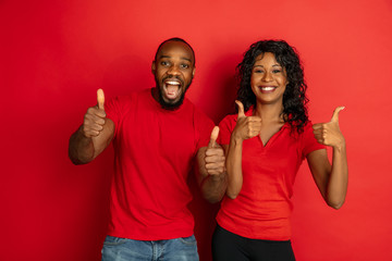 Young emotional african-american man and woman in bright casual clothes posing on red background. Concept of human emotions, facial expession, relations, ad. Couple showing thumbs up and smiling.