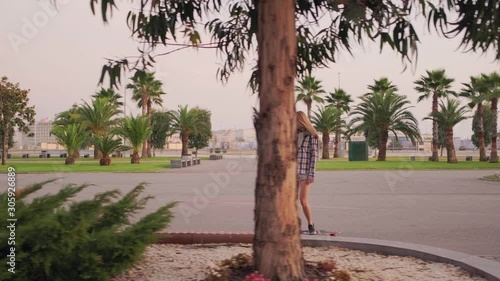 Slow motion, a young sophisticated Woman in a shirt and denim shorts rides a Skateboard through the Park among the palm trees. Hair develops at speed. Trackng Shoit. photo