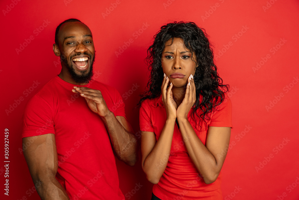 Young emotional african-american man and woman in bright casual clothes posing on red background. Concept of human emotions, facial expession, relations, ad. She upset and sad, he happy. Cute couple. Young emotional african-american man and woman in bright casual clothes posing on red background. Concept of human emotions, facial expession, relations, ad. She upset and sad, he happy. Cute couple.