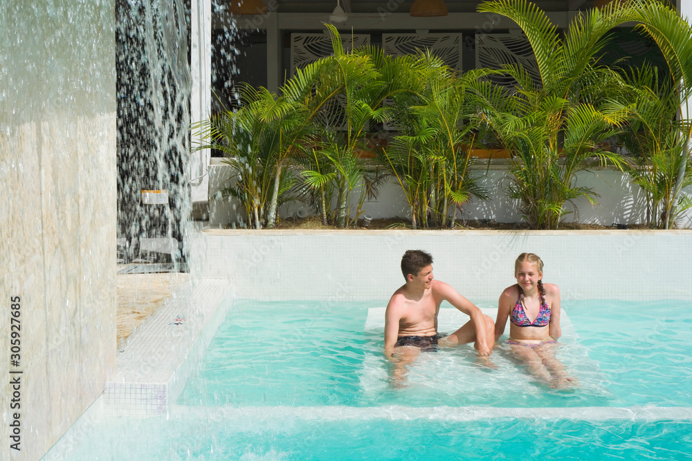 Caucasian teenagers sitting on the bed and relaxing in swimming pool in luxury hotel, Punta Cana, Dominican Republic. Artificial waterfall on the left. Summer vacation concept