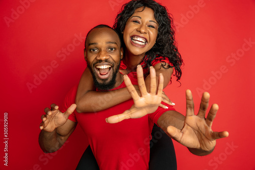 Young emotional african-american man and woman in bright casual clothes posing on red background. Concept of human emotions, facial expession, relations, ad. Cute couple hugging and laughting. photo