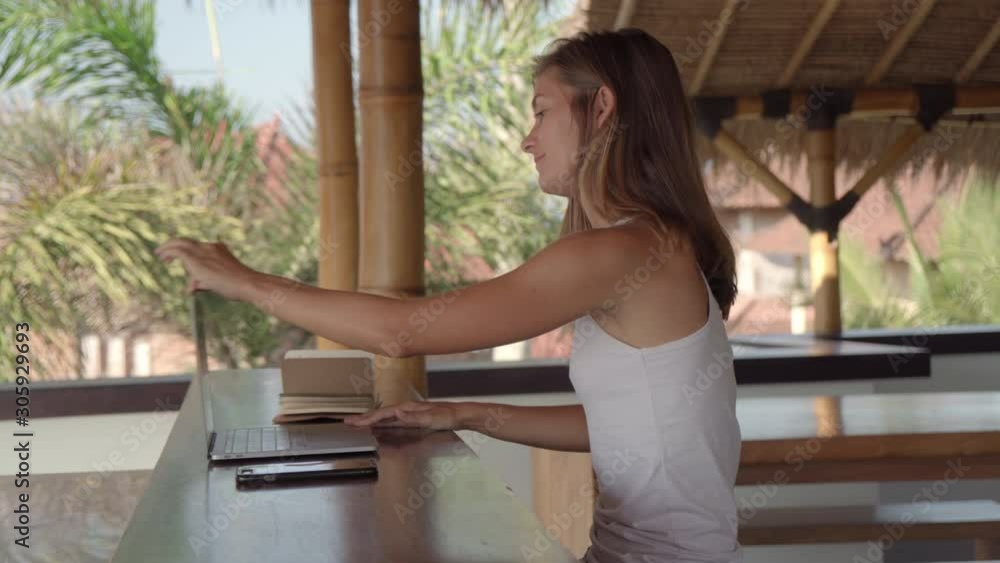 Sequence of shots of Caucasian woman walking to her workplace in thatched tropical patio, sitting at desk, using laptop, talking on phone and writing in notepad while working remotely on vacation Sequence of shots of Caucasian woman walking to her workplace in thatched tropical patio, sitting at desk, using laptop, talking on phone and writing in notepad while working remotely on vacation