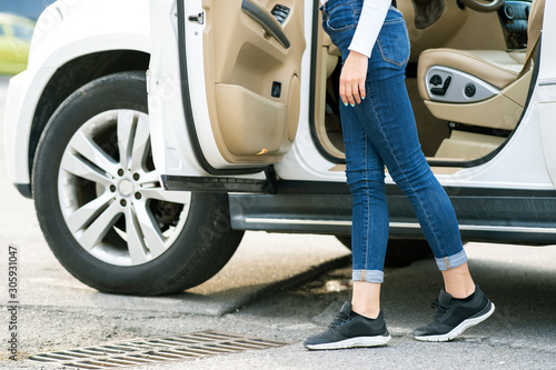 A woman in jeans and sneakers standing near a big all terrain car outdoors. Driver girl in casual clothes outside her vehicle. © bilanol