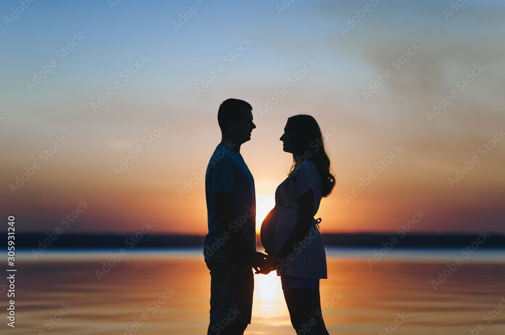 Loving man holds the hand of a pregnant woman at sunset, against the background of the sea, river, standing on the pier. Portrait of beautiful newlyweds expecting a baby. Photography, concept.