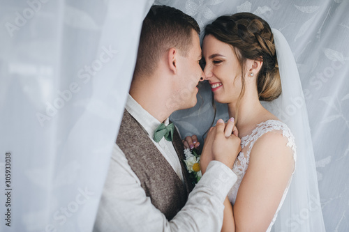 Wedding portrait of beautiful, lovers, smiling newlyweds in curtains on a background of fabric. Stylish groom in a suit hugs a cute bride in a white dress holding her hand. Photography and concept.