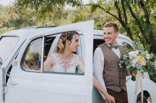 Loving newlyweds are standing, holding hands, near an old white retro car on nature in summer. Wedding portrait of a stylish, smiling groom and a cute bride with curly hair. Photography and concept.