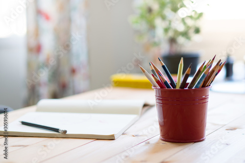 Close up of red tin bucket full of colorful color pencils and opened notebook with pencil on wooden table. Neat workspace at home. Early child development.