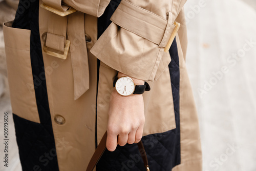 street style fashion details. close up, young fashion blogger wearing autumn trench coat and a white and golden black analog wrist watch. checking the time, holding a beautiful brown leather purse.