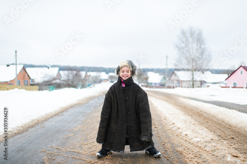 Little cheerful girl in oversized padded jacket standing on snowy road in winter day.