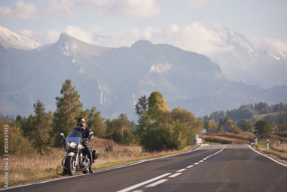 Young bearded motorcyclist in black leather jacket sitting on bike on country roadside on blurred background of green rural landscape, distant white sunny mountain peaks and bright sky.
