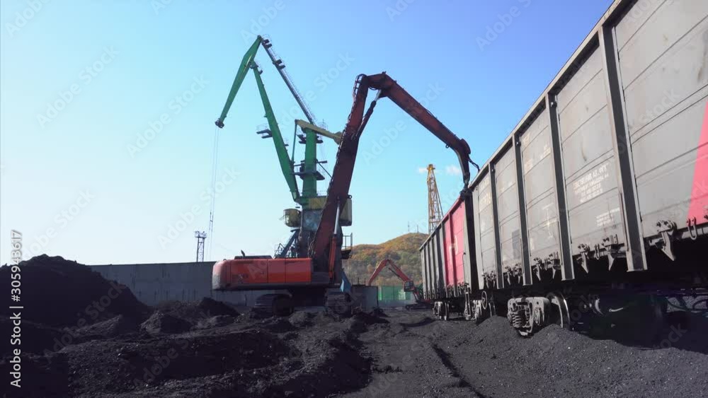 Shooting from below. A large, powerful excavator with a bucket loads black anthracite coal into gray-red railway cars in the seaport of a coal company.