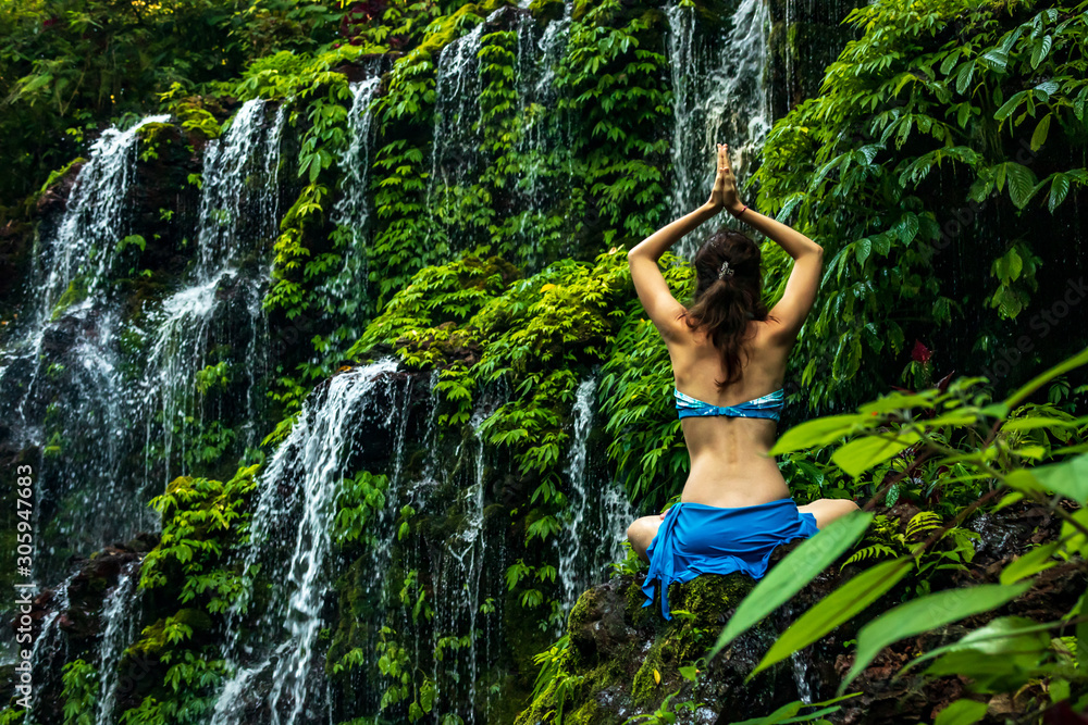 Woman sitting on the rock, practicing yoga. Young woman raising arms with namaste mudra near waterfall. Banyu Wana Amertha waterfall, Bali. View from back. Woman sitting on the rock, practicing yoga. Young woman raising arms with namaste mudra near waterfall. Banyu Wana Amertha waterfall, Bali. View from back.