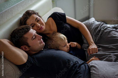 Happy family at home bedroom lies with kid on the bed together under the blanket. People wearing black t-shirt, shorts and baby bodysuit.