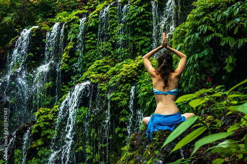 Woman sitting on the rock  practicing yoga. Young woman raising arms with namaste mudra near waterfall. Banyu Wana Amertha waterfall  Bali. View from back.