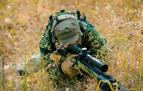 Airsoft man in uniform lay in long grass with sniper rifle. Soldier aims at the sight