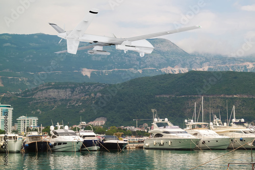 unmanned RC military drone flies above yachts and boats in harbor with mountains