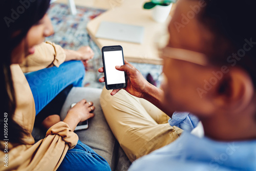 Cropped view of man holding mobile phone with mock up screen showing photos to his girlfriend sitting on sofa at home, back view of hipster guy holding smartphone with blank copy space screen