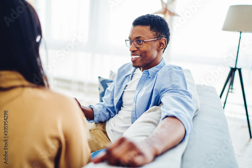 Cheerful African American man talking with his girlfriend sitting on sofa in modern apartment together, dark skinned couple in love communicating with each other enjoying relationships at home