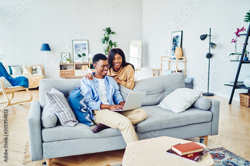Cheerful african american wifie looking at her husband working on freelance via laptop computer at home interior, dark skinned romantic couple booking tickets for trip vacation together via netbook. #305954852