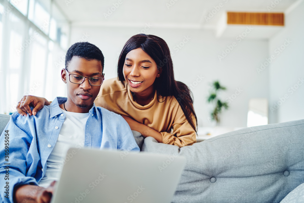 Smiling african american woman looking at freelance job of husband sitting with laptop computer at sofa in living room, positive dark skinned couple in love making shopping together in web store. Smiling african american woman looking at freelance job of husband sitting with laptop computer at sofa in living room, positive dark skinned couple in love making shopping together in web store.