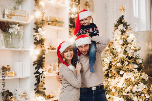 Portrait of cheerful young family in Santa hats celebrating New Year at home. They standing by beautifully decorated Christmas tree with balls and garland. Looking at camera. Snowfall.