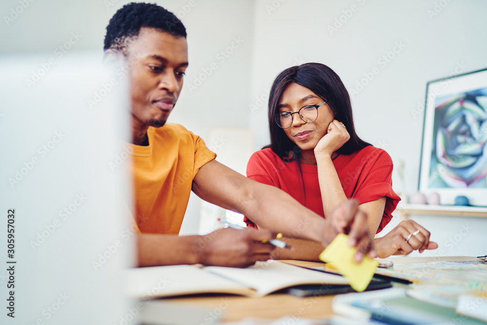Smiling African American woman looking how her husband working on freelance at home, positive dark skinned female satisfies with cooperation with friend sitting at desktop learning and make notes Smiling African American woman looking how her husband working on freelance at home, positive dark skinned female satisfies with cooperation with friend sitting at desktop learning and make notes