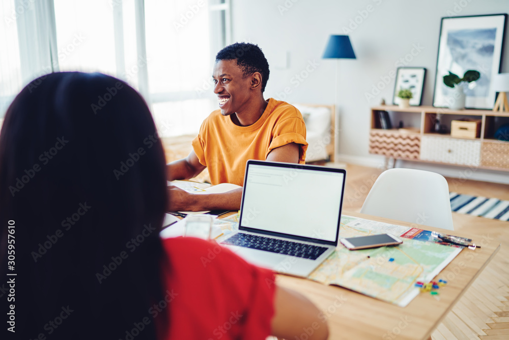 Cropped image with back view of woman sitting at table near cheerful african american male student, smiling dark skinned hipster guy satisfied with communication during planning trip using netbook. Cropped image with back view of woman sitting at table near cheerful african american male student, smiling dark skinned hipster guy satisfied with communication during planning trip using netbook.