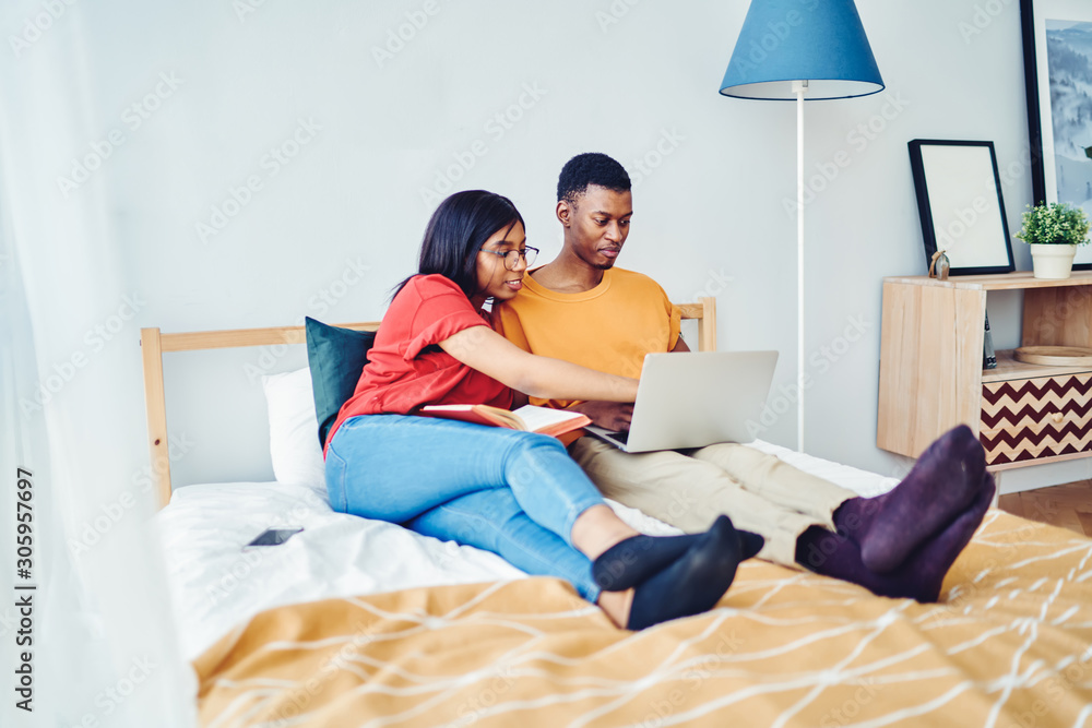 African american hipster girl using app on boyfriend's laptop computer for watching online videos while resting at bed together, couple browse web page on netbook making booking together at home.