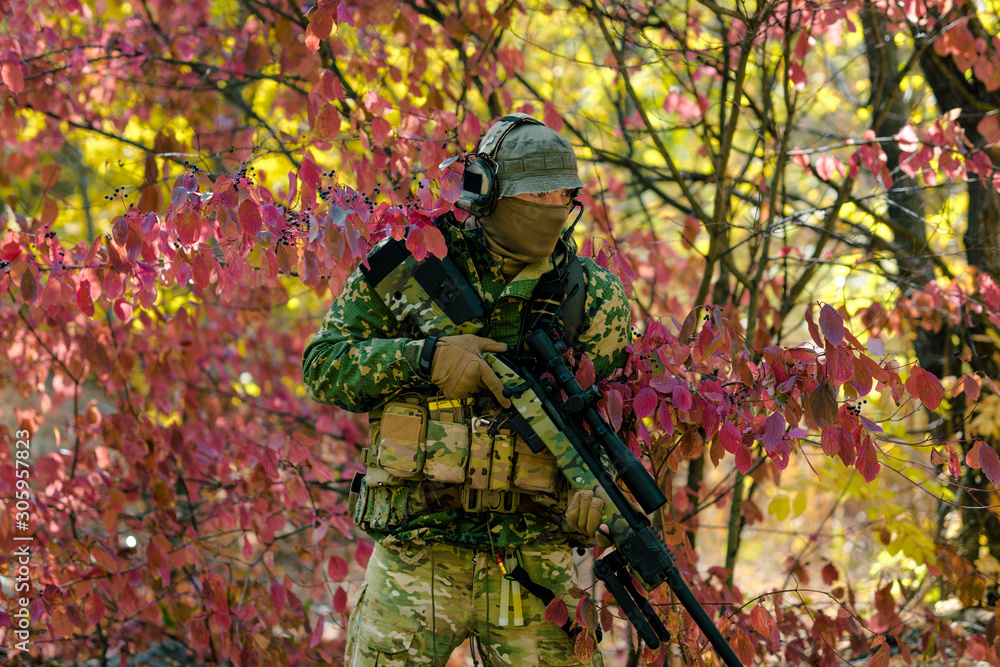 Naklejka premium Man in green uniform with sniper rifle. Airsoft soldier stand in red bush. Horizontal photo