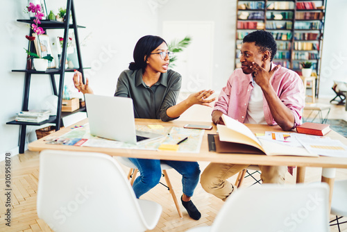 Emotional dark skinned woman gesture while talking to male colleague sitting together at table with laptop computer, young african american students having productive discussion about project.