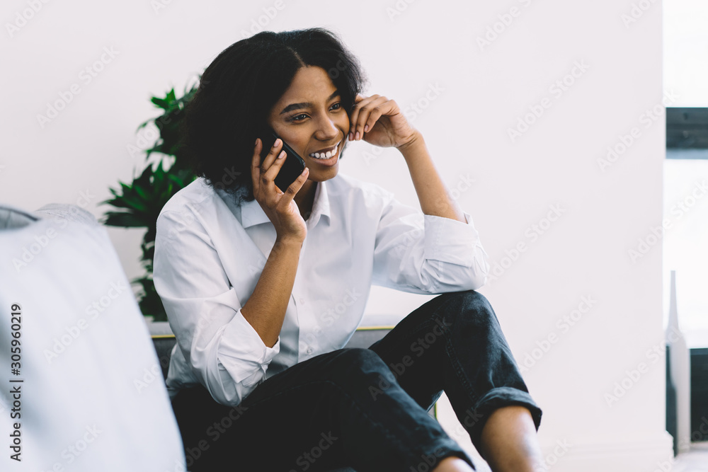 Pretty young woman with dark skin smiling and talking on smartphone with friend resting on comfortable sofa in modern apartment.Positive african american hipster girl calling on mobile phone at home Pretty young woman with dark skin smiling and talking on smartphone with friend resting on comfortable sofa in modern apartment.Positive african american hipster girl calling on mobile phone at home