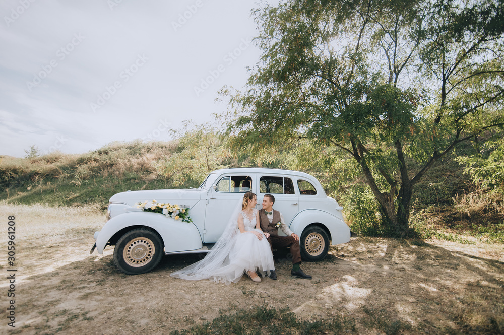 Beautiful newlyweds are sitting and hugging near an old retro car and summer nature. Wedding portrait of a stylish, smiling groom and lovely bride with curly hair. Photography and concept.