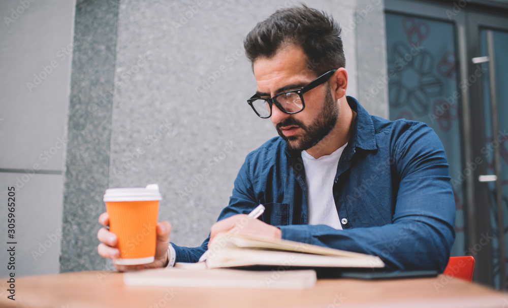 Concentrated male student in classic spectacles for vision correction sitting at street cafeteria and learning preparing to university exams, bearded man making memo notes in knowledge textbook Concentrated male student in classic spectacles for vision correction sitting at street cafeteria and learning preparing to university exams, bearded man making memo notes in knowledge textbook