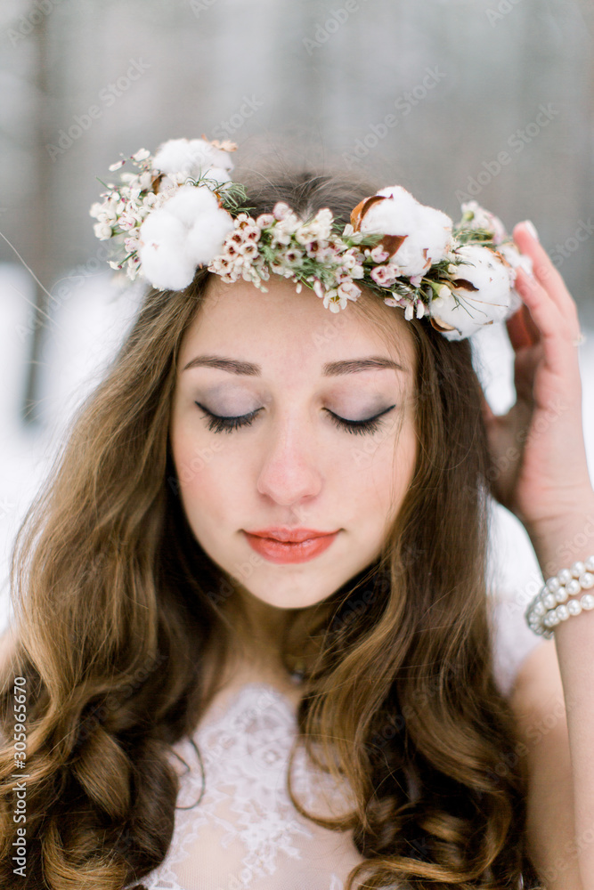 Beautiful bride with curly hair, touching her floral head band wreath, made from flowers and cotton, standing outdoors, on the background of winter forest Beautiful bride with curly hair, touching her floral head band wreath, made from flowers and cotton, standing outdoors, on the background of winter forest