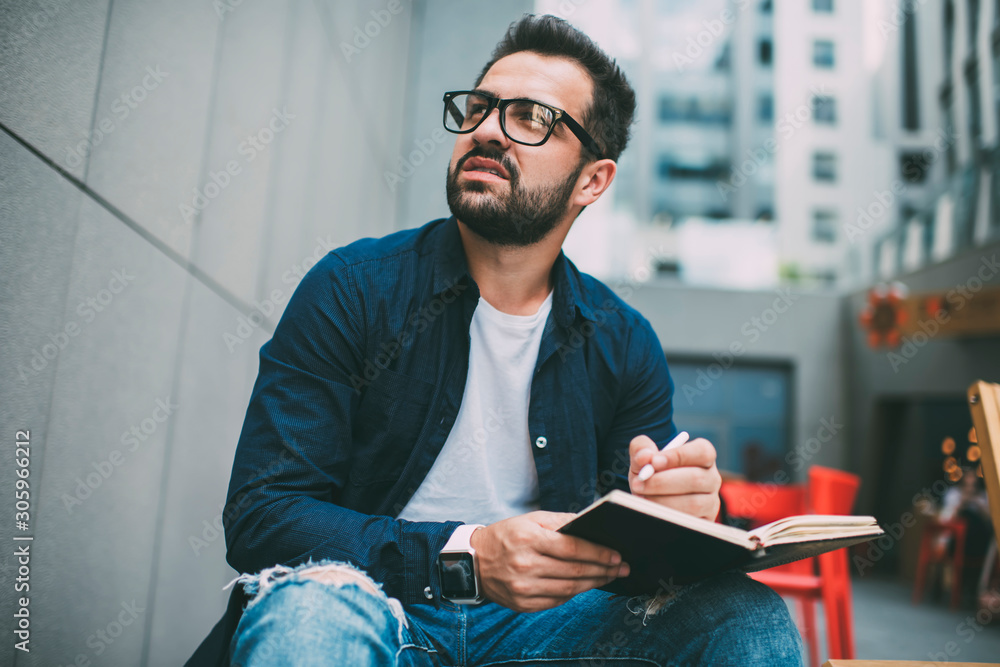Pensive hipster guy in optical spectacles for vision correction thoughtful looking away while planning week schedule with knowledge notepad, contemplative clever man thinking on idea for course work Pensive hipster guy in optical spectacles for vision correction thoughtful looking away while planning week schedule with knowledge notepad, contemplative clever man thinking on idea for course work