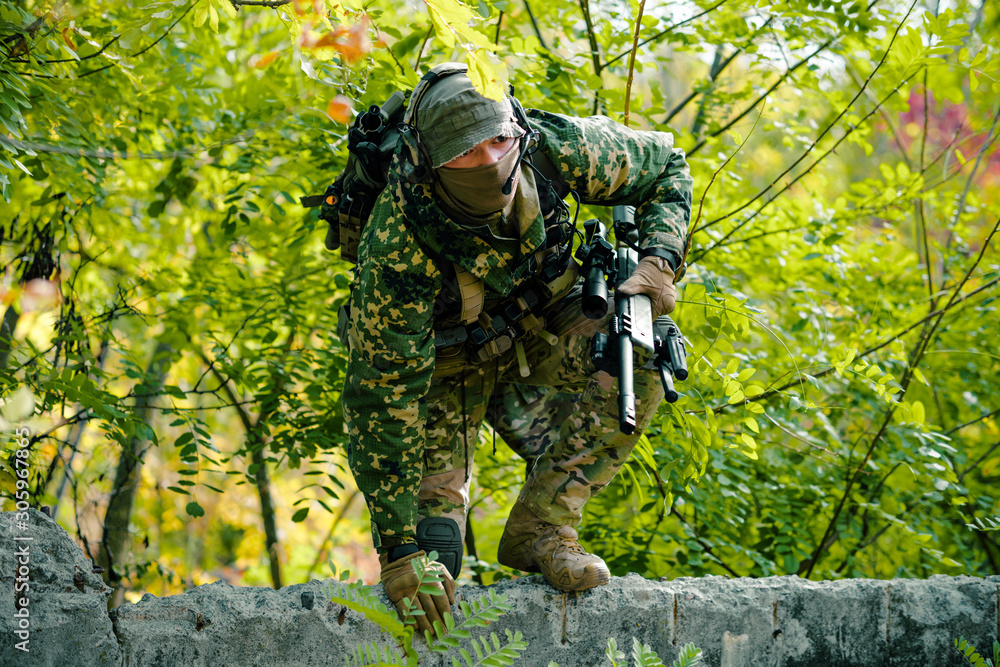 Man in uniform climbs over the wall with sniper rifle on green forest background