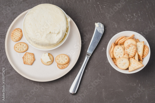 Cheese and cracker in a white bowl with a knife on a dark stone background. Top view.