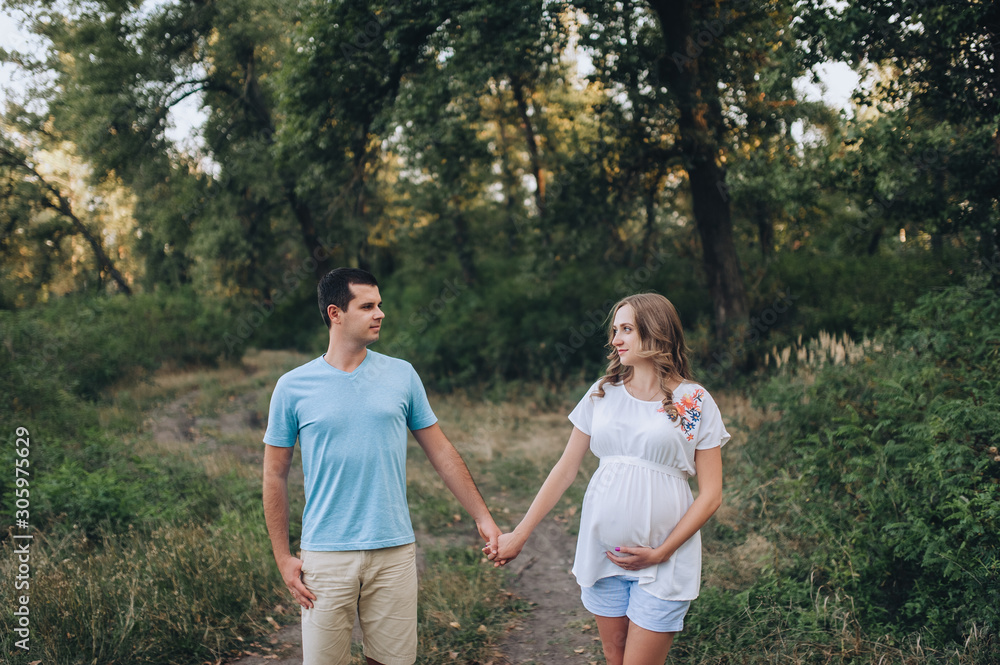 Fototapeta premium Portrait of a young family expecting a baby. A loving man and a beautiful pregnant blond woman holding hands at sunset in the forest. Pregnancy picture, concept.