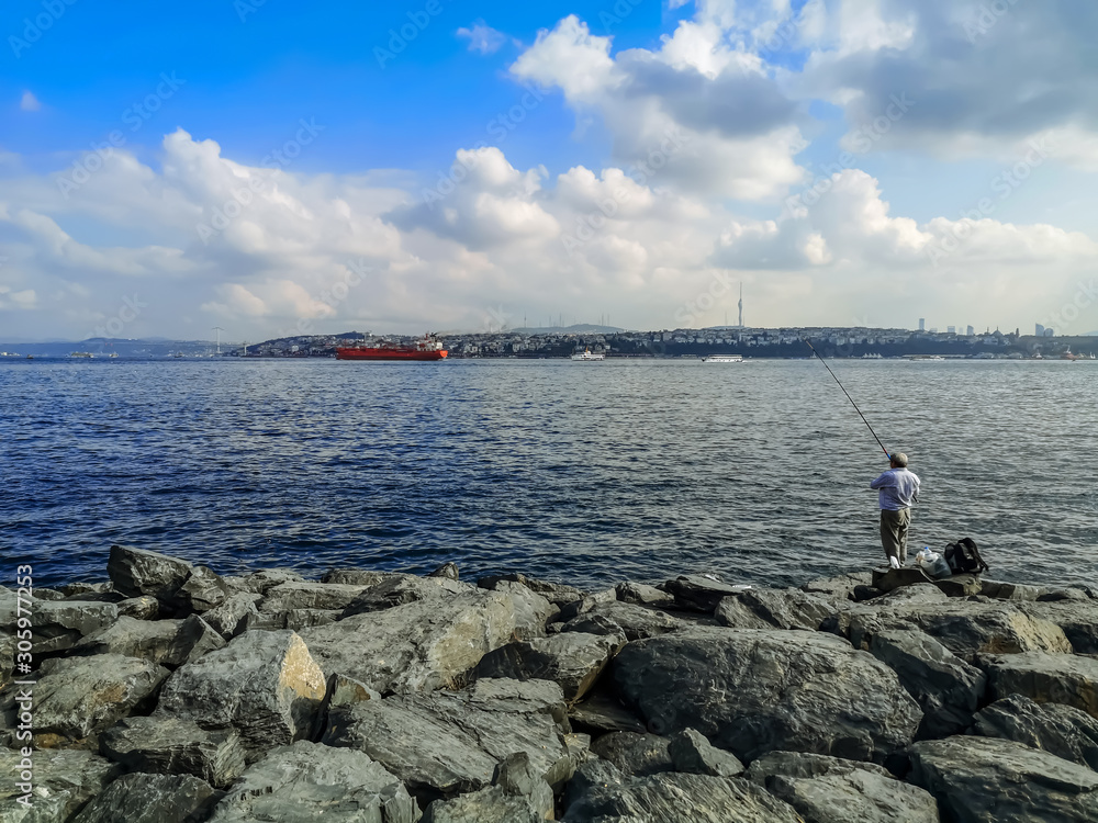 Fisherman fishes in the Sea of Marmara on the promenade in Istanbul, Turkey. Large boulders on the banks of the Bosphorus, a large red tanker at sea, and a cityscape on the other side of the bay Fisherman fishes in the Sea of Marmara on the promenade in Istanbul, Turkey. Large boulders on the banks of the Bosphorus, a large red tanker at sea, and a cityscape on the other side of the bay