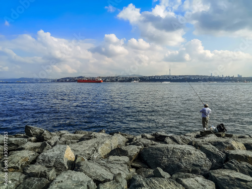 Fisherman fishes in the Sea of ​​Marmara on the promenade in Istanbul, Turkey. Large boulders on the banks of the Bosphorus, a large red tanker at sea, and a cityscape on the other side of the bay