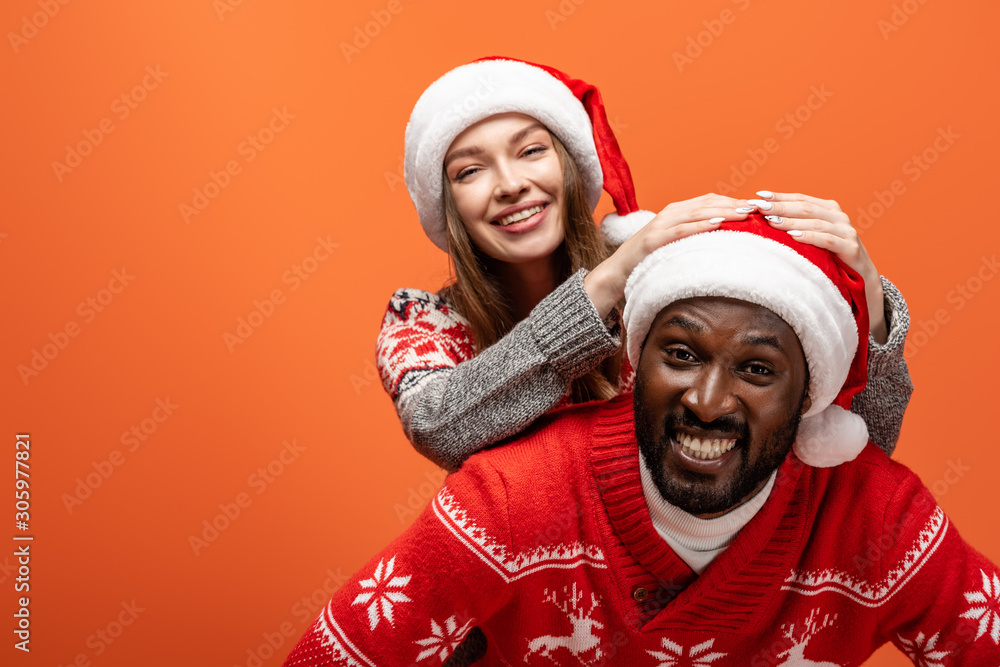 Fototapeta premium happy interracial couple in santa hats and Christmas sweaters on orange background