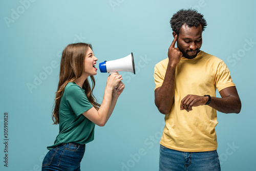 angry girl screaming in megaphone at african american boyfriend looking at wristwatch on blue background