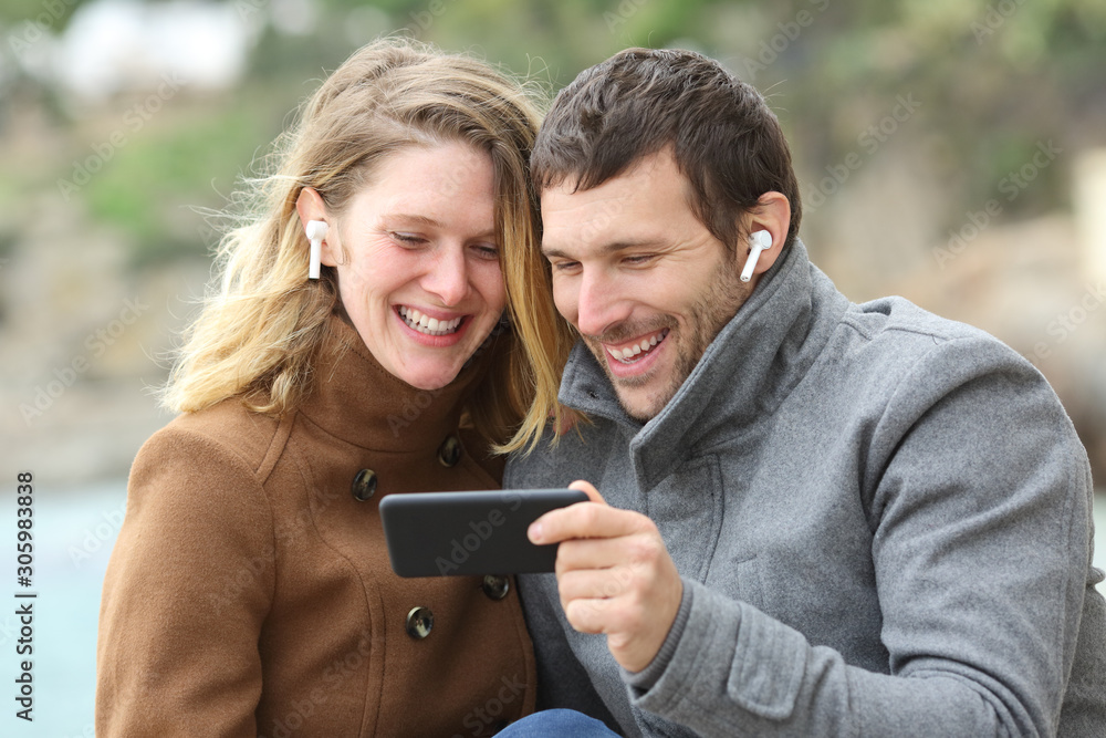 Happy couple watching videos sharing earbuds Happy couple watching videos sharing earbuds